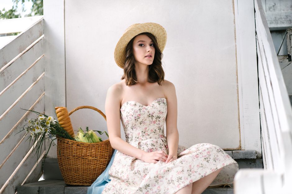 A woman in a floral dress and straw hat sits on stairs with a basket of groceries.