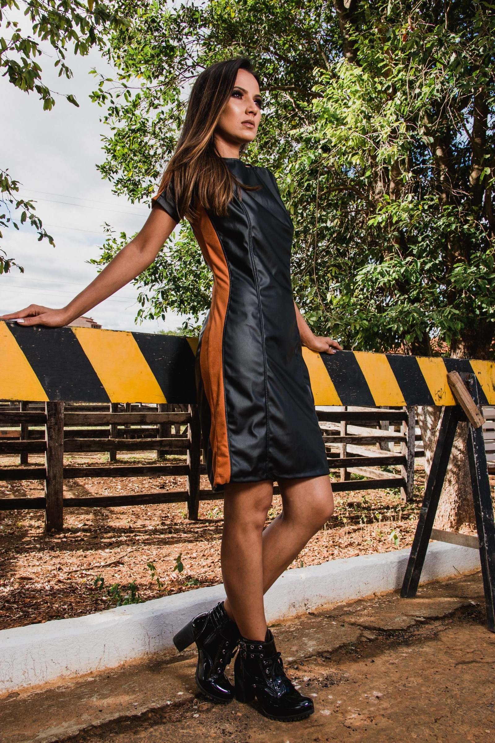 Woman wearing an above-knee dark dress and black heeled boots beside a rustic fence