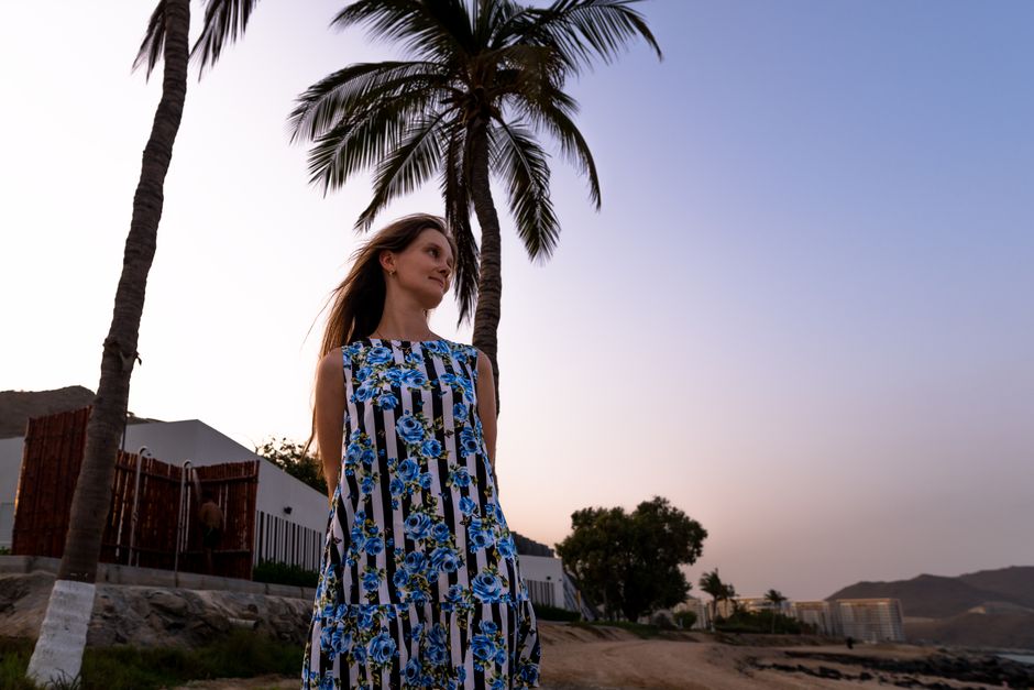 Woman in floral dress stands by palm trees at sunset at Хор-Факкан beach, UAE.